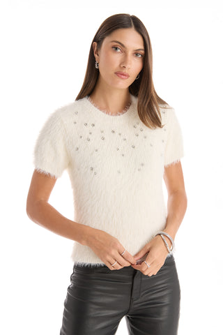 A woman with long brown hair models the Angel Fluffy Sweater Top - White, a soft short-sleeve crewneck sweater with small embellishments, styled with black leather pants and silver jewelry against a white background.