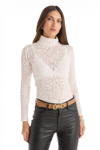 A woman stands against a white background wearing the Alina Rose Turtleneck Top - Pearl, a slim-fitting, rose motif lace long-sleeve over a white bra, styled with black leather pants and a tan belt.