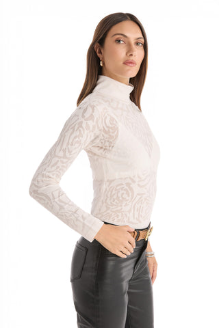 A woman with long brown hair wears the Alina Rose Turtleneck Top - Pearl, a slim-fitting, white patterned rose lace top, paired with black leather pants. She stands sideways and looks neutrally at the camera against a white background.