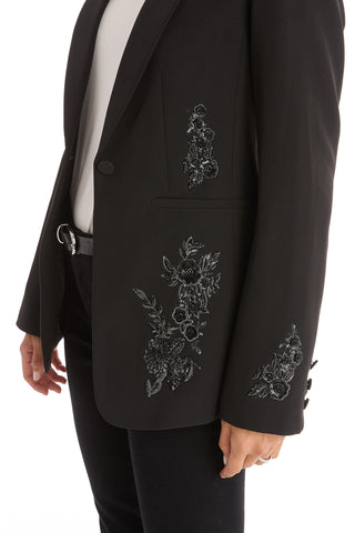 Wearing the London Floral Sequin Blazer - Black, featuring black floral embroidery on the pocket and sleeve, styled with a white top and black pants for an effortlessly chic look.