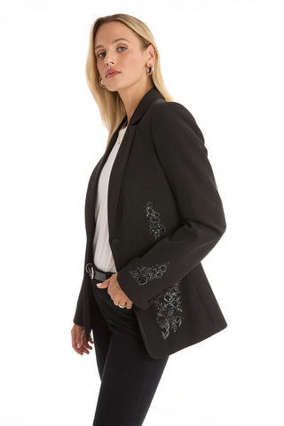 A woman with long blonde hair models the London Floral Sequin Blazer - Black over a white blouse and black pants, standing sideways and looking confidently at the camera against a plain white background.