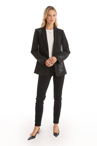 A woman with long blonde hair and a neutral expression stands against a white background, wearing the London Floral Sequin Blazer in black over a white top, paired with black pants and heels. Her hands are clasped in front.
