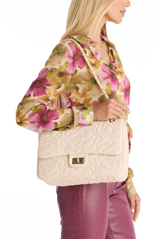 A woman in a floral blouse and pink pants holds the Alessia Rose Embossed Leather Bag in Pearl, a quilted purse with a gold chain strap and clasp, shown from the side against a white background.