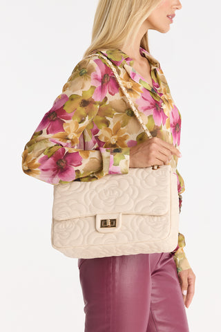 A woman with long blonde hair holds the Alessia Rose Embossed Leather Bag in Pearl—a cream quilted handbag with a gold chain strap and subtle floral pattern—pairing it with a floral blouse and mauve pants. Captured from shoulders to mid-thigh.