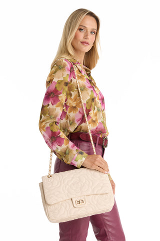 A woman in a floral blouse and mauve pants holds the Alessia Rose Embossed Leather Bag in Pearl, a quilted cream handbag with a chain strap, standing against a white background.