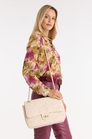 A blonde woman in a floral blouse and pink leather pants holds the Alessia Rose Embossed Leather Bag - Pearl with a chain strap, standing against a plain white background and glancing to the side.