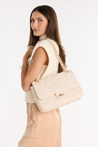 A woman with long brown hair wears a sleeveless white knit top and beige pants, posing with the Alessia Rose Embossed Leather Bag - Pearl, featuring a gold chain strap, over her shoulder against a white background.