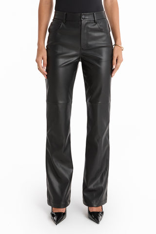 A person models the Jackie Leather Pant - Black, styled with black pointed-toe heels against a white background. The image focuses on the lower torso and legs, showcasing the pant's sleek faux leather design.