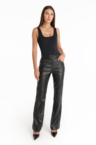A woman stands against a white background wearing the Jackie Leather Pant - Black, styled with a black sleeveless top and black pointed-toe shoes, one hand in her pocket and the other relaxed by her side.