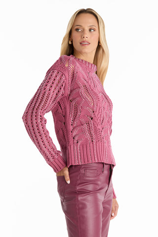 A woman with long blonde hair wears the Elea Crystal Knit Sweater in Sangria, paired with matching pink leather pants. She stands slightly turned to the side, looking over her shoulder against a white background.