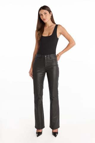 A woman faces forward in the Loren Bodysuit - Black, paired with high-waisted black leather pants and black pointed-toe heels, against a plain white background.