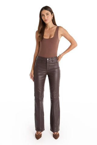 A woman with long brown hair and a neutral expression wears the Loren Bodysuit - Espresso, high-waisted dark brown leather pants, pointed-toe brown heels, and gold jewelry against a white background.