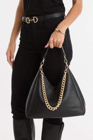 Wearing black pants, a black top, and a black belt, a person holds the Tribeca Chain Leather Bag - Black with gold chain detail; only their lower torso, right arm, and upper legs are shown.