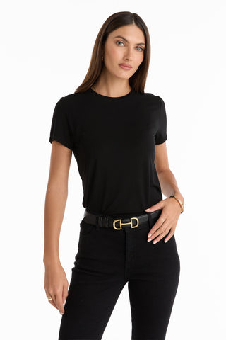 A woman with long brown hair poses confidently against a white background, wearing the Amaya Ultra-Soft T-Shirt in black, paired with black pants and a black belt featuring a gold buckle.