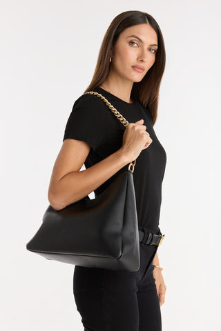 A woman with long brown hair, dressed in a black t-shirt and pants, poses against a white background carrying the Tribeca Chain Leather Bag - Black over her shoulder, showcasing its unique gold chain detail.