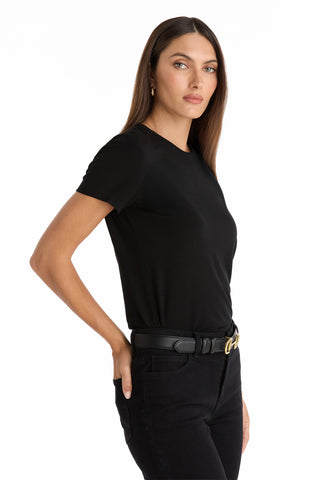 A woman with long brown hair, dressed in the Amaya Ultra-Soft T-Shirt in black and matching pants with a black belt, stands against a white background, angled slightly to the side with one hand in her pocket.