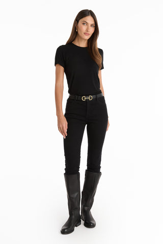 A woman with straight brown hair and a neutral expression stands against a white background wearing the Amaya Ultra-Soft T-Shirt in black, paired with black pants, a belt, and knee-high black boots.