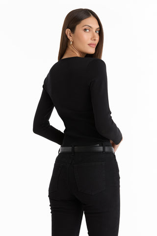 A woman with long brown hair wears the Edie Knit Top in black, paired with black jeans and a black belt. She stands turned slightly away, glancing over her shoulder against a white background.