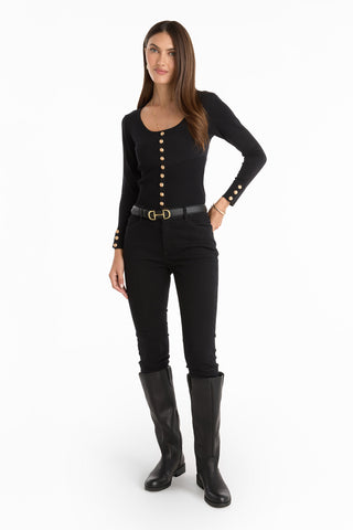 A woman with long brown hair poses against a white background wearing the Edie Knit Top - Black, styled with black pants, a gold-buckled belt, and knee-high boots. She looks relaxed and elegant.