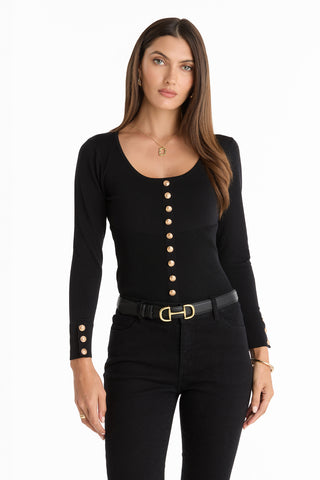 A woman with long brown hair wears the Edie Knit Top in black, paired with black pants and a black belt with a gold buckle, standing against a plain white background.