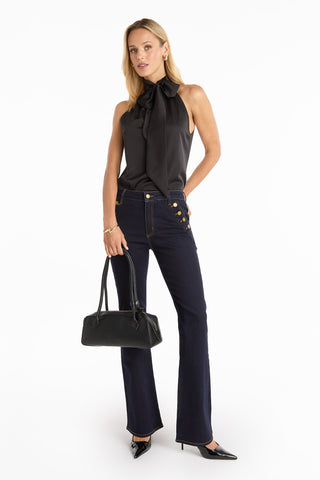 A woman stands against a white background wearing a sleeveless black bow blouse, dark high-waisted jeans, black pointed-toe heels, and holding the Reese Leather Bag - Black with gold hardware.