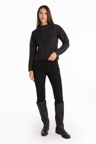 A woman with straight brown hair and a neutral expression stands against a white background wearing the Elea Crystal Knit Sweater - Black, paired with black skinny pants and knee-high boots, with one hand on her hip.