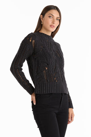 A woman stands against a white background, looking slightly to the side, wearing the Elea Crystal Knit Sweater in black and matching black pants.