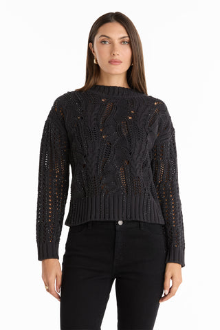 A woman with straight brown hair and a neutral expression faces forward, wearing the Elea Crystal Knit Sweater in black with a textured openwork pattern, paired with black pants, against a plain white background.