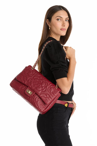 A woman with long brown hair in a black outfit holds the Alessia Rose Embossed Leather Bag - Merlot, a large quilted red handbag with a gold chain strap and subtle rose-embossed design, standing against a plain white background.