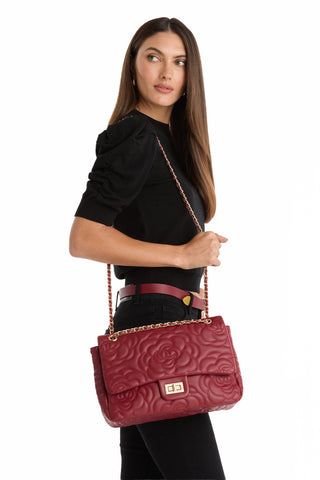 Woman with long brown hair in a black top and pants holds the Alessia Rose Embossed Leather Bag - Merlot, featuring a chain strap over her shoulder, while looking to the side against a white background.
