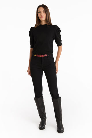 A woman stands against a white background wearing the Sylvie Knit Top - Black, styled with black jeans, a pink belt with a gold buckle, and knee-high black boots—an essential look for your fall wardrobe.