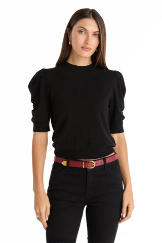 A woman with long brown hair wears the Sylvie Knit Top - Black, featuring puffed shoulders, styled with black pants and a bold red belt. She stands against a plain white background, showcasing an effortless fall wardrobe update.