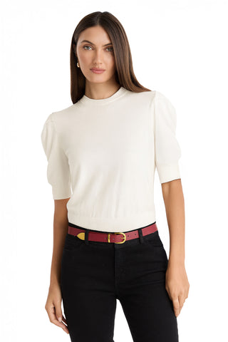 A woman with straight brown hair wears the Sylvie Knit Top in Pearl, paired with black pants and a red belt with a gold buckle. She stands against a plain white background, looking at the camera with a neutral expression.