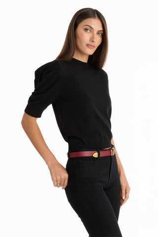 A woman with straight brown hair wears the Sylvie Knit Top - Black, featuring a fitted crewneck and elbow-length sleeves, paired with black jeans and a red belt against a white background—an ideal elevated fall look.