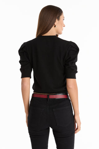 A woman with long brown hair, dressed in the Sylvie Knit Top - Black with puffed shoulders, black jeans, and a red belt, stands facing away from the camera against a white background—a chic choice for any Fall wardrobe.