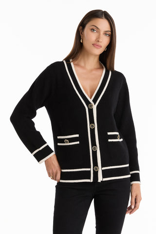 A woman models the Devin Rose Contrast Cardigan in black, featuring white trim and gold buttons, styled with black pants and a hand in her pocket against a plain white background.