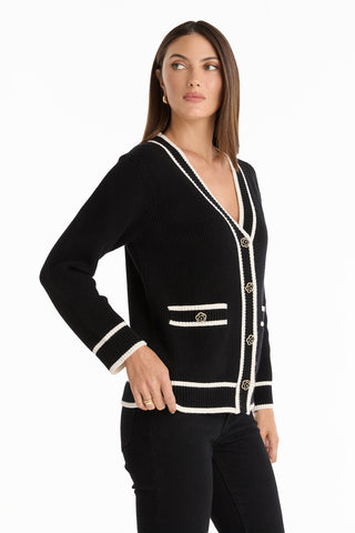 A woman models the Devin Rose Contrast Cardigan - Black, featuring white trim, gold buttons, and front pockets, standing sideways with her hand on her hip against a plain white background.
