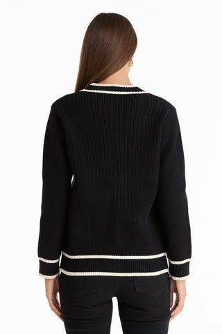 A person with long brown hair is seen from behind wearing the Devin Rose Contrast Cardigan - Black, featuring white trim on the collar, cuffs, and hem, paired with black pants against a white background.