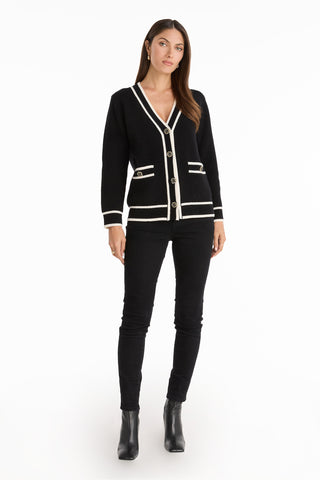 A woman faces forward against a plain white background, wearing the Devin Rose Contrast Cardigan in black with white trim, black skinny pants, and black ankle boots.