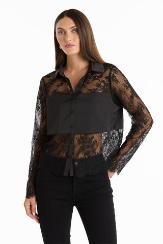A woman poses against a white background, wearing the Vienna Lace Blouse - Black with black lace long sleeves, a solid black panel on the chest and collar, paired with sleek black pants.
