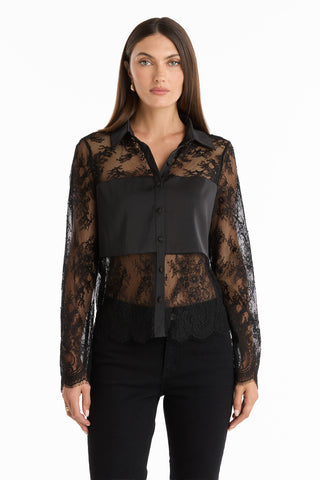 A woman with straight brown hair and a neutral expression stands facing forward in the Vienna Lace Blouse - Black, an elegant lace top with a faux leather chest panel, paired with black pants against a white background.