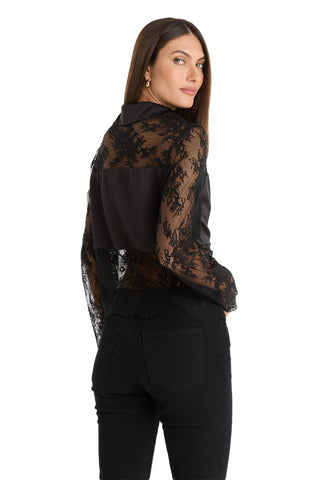A woman with long brown hair stands facing away, turning her head slightly toward the camera. She wears the Vienna Lace Blouse - Black featuring sheer sleeves and back, paired with black jeans. The background is white.