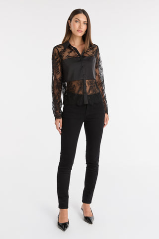 A woman with straight brown hair and a neutral expression stands against a white background, wearing the Vienna Lace Blouse - Black paired with black skinny pants and pointed-toe heels. Her arms are relaxed at her sides.