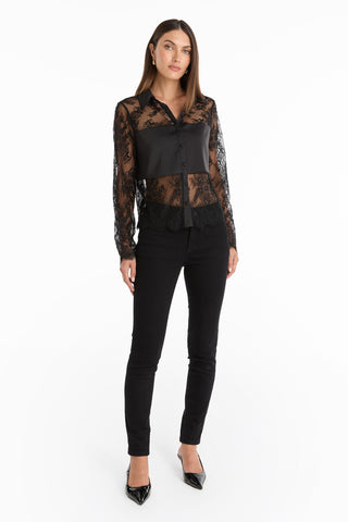 A woman with straight dark hair and a neutral expression stands against a white background, wearing the Vienna Lace Blouse - Black, paired with black pants and black pointed-toe heels. Her arms are relaxed at her sides.