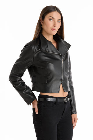 A woman with long brown hair wears the Katalina Moto Jacket - Black with zipper cuffs, paired with black jeans and a black belt. She stands against a white background, one hand in her pocket.