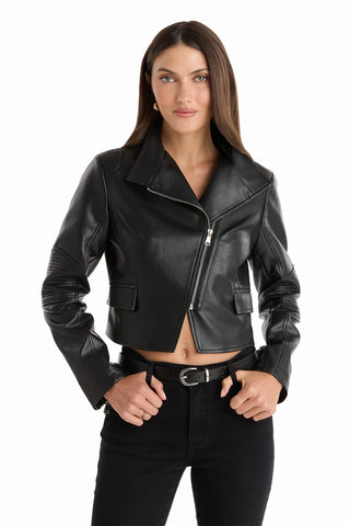 A woman with straight brown hair wears the black Katalina Moto Jacket with zipper cuffs and black belted jeans, standing against a white background with her hands on her hips.