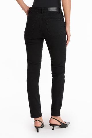 A person stands facing away, wearing the Layla Skinny Jean - Black. These high-waisted black skinny jeans feature back pockets, a leather patch label on the waistband, and are paired with black heeled sandals against a plain white background.