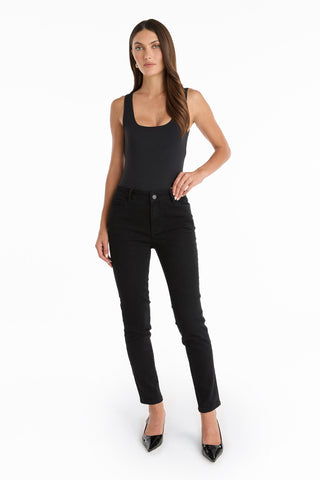 A woman stands against a white background wearing a black tank top, the Layla Skinny Jean - Black, and black high-heeled shoes, with her hair down and one hand on her hip.