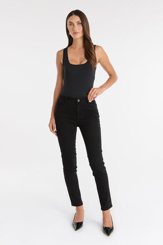 A woman with long brown hair and a neutral expression stands against a white background wearing the Layla Skinny Jean - Black, a sleeveless black bodysuit, and black pointed-toe heels.