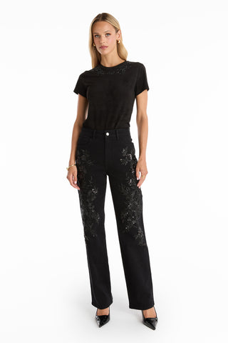 A woman with straight blonde hair stands against a white background wearing the Amaya Floral Sequin T-Shirt in black, paired with high-waisted black jeans featuring embroidery and black pointed-toe heels.